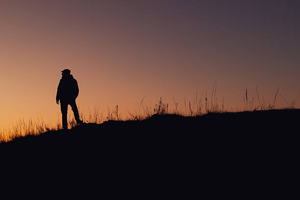 A person is standing on a hill at sunset. The sky is a beautiful mix of orange and purple hues. The person is wearing a backpack and he is enjoying the view