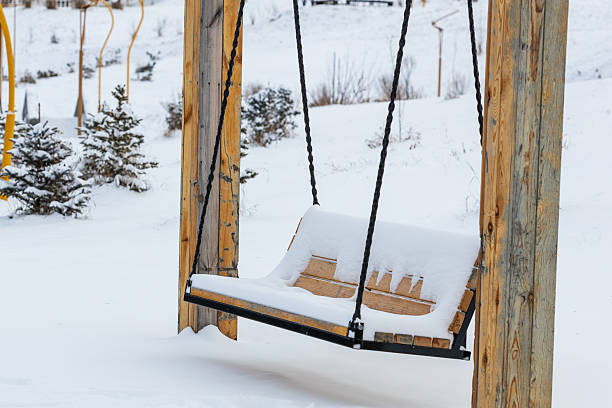 A wooden swing is covered in snow. The snow is piled up on the seat and the chains. The snow is white and fluffy