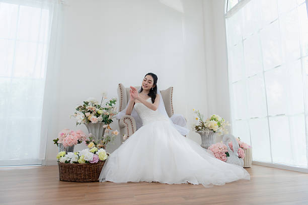 Wedding, Bride in beautiful dress sitting on sofa indoors in white studio interior, Young attractive Asian woman Smile brightly.