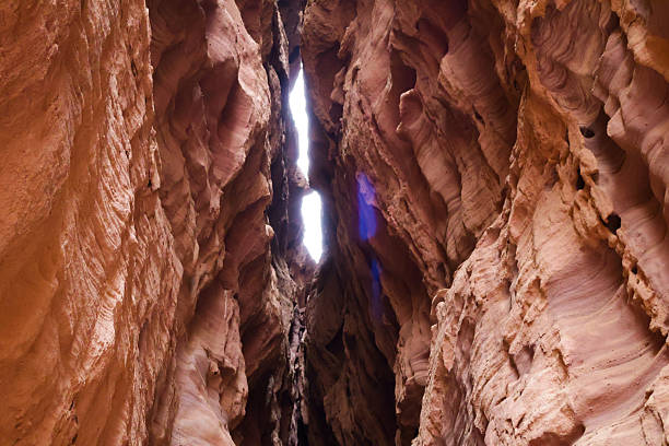 Dramatic red sandstone slot canyon with high, rugged walls and a bright gap at the top. Warm tones, textured surfaces, and a sense of awe and adventure in the tight canyon passage.