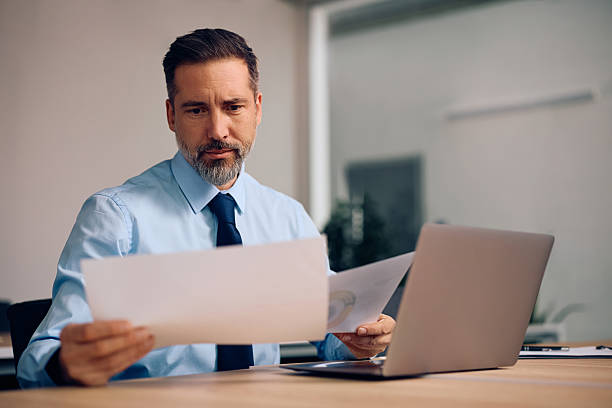 istock Mature businessman going through paperwork while using laptop and working in the office. 2254628470