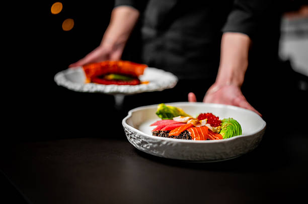A chef presents an elegant, fresh poke bowl featuring salmon, avocado, roe, and tuna in a fine white bowl.