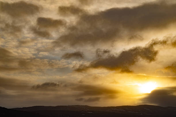 Golden sunrise emerging through layered clouds above a distant landscape. Warm sunlight contrasts with soft, textured cloud formations, creating a calm yet powerful atmosphere of transition, renewal, and natural beauty. Suitable for themes related to hope, change, weather, and new beginnings.