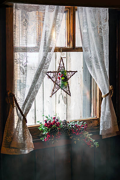 Cozy rustic interior featuring a wooden window with sheer lace curtains, a hanging Christmas star ornament, and festive greenery with red berries, illuminated by soft natural winter light