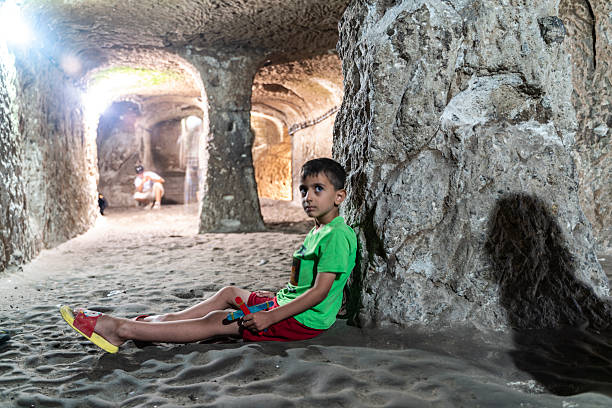 A young boy sits quietly against a carved stone wall inside the ancient tunnels of Derinkuyu Underground City in Cappadocia, Turkey. The historic subterranean environment, shaped from volcanic rock centuries ago, creates a powerful contrast between childhood innocence and humanity’s deep past. This image conveys themes of cultural heritage, travel, history, curiosity, and exploration within one of the world’s most fascinating underground settlements.