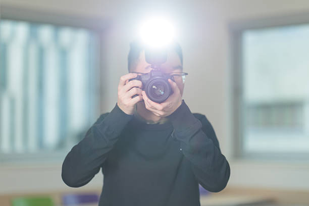 A Chinese student holds a camera with flash pointed directly at viewer, ideal for photography education, media studies, or creative arts concepts.