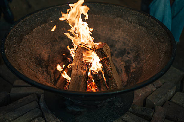 Atmospheric close-up shot of burning wood logs in a rustic metal fire pit during nighttime. Bright orange flames and glowing embers create a warm, cozy ambiance against the dark background. The circular metal pit contains burning firewood with dancing flames and rising smoke, perfect for outdoor entertainment and warmth.