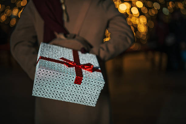 Person holding a shimmering silver gift box wrapped with a red ribbon and bow behind their back, preparing a thoughtful surprise during a festive celebration with bokeh lights