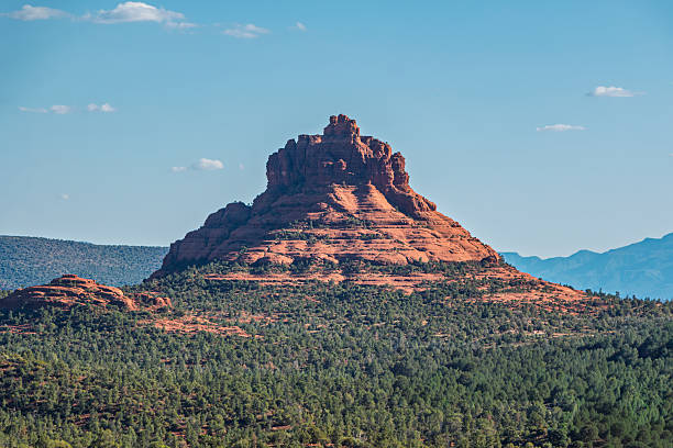A landmark red rock formation is centered in the image, surrounded by a landscape of trees and shrubs under a blue sky. Sedona, Arizona.
