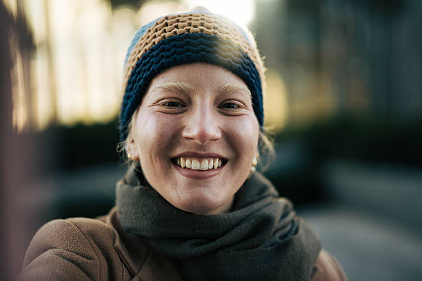 Young adult woman wearing a knitted hat and scarf, smiling sincerely at the camera while taking a selfie outdoors in an urban setting with bokeh background
