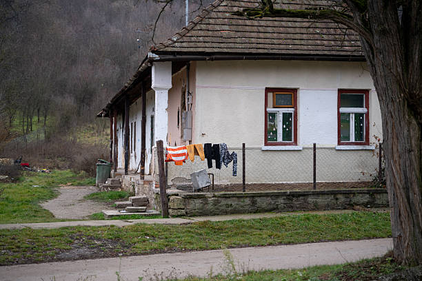 A weathered countryside house with a tiled roof stands in a quiet rural setting. Colorful clothes hang on the fence to dry, adding a touch of everyday life to the rustic and slightly run-down environment. The background features bare trees and rolling hills, suggesting a cold season.