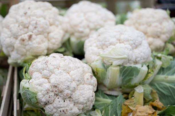 Close-up of multiple fresh cauliflower heads displayed on a market stall, with green leaves surrounding the white florets. The earthy texture and slight imperfections on the surface emphasize their organic and natural state, evoking a rustic and healthy food atmosphere.