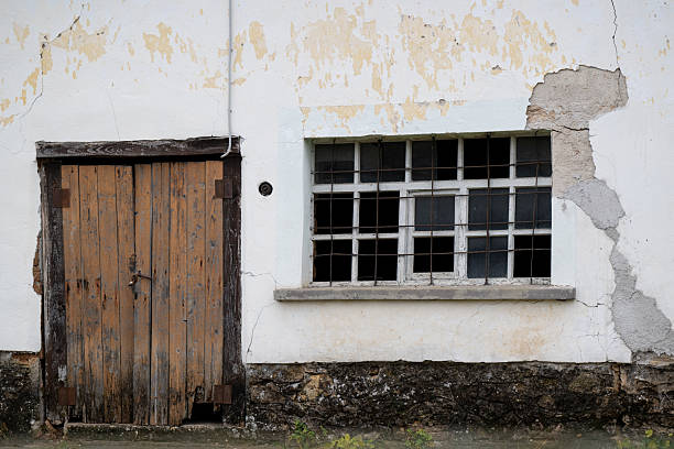 Front view of a deteriorating building facade featuring a worn wooden door and a damaged, grid-framed window. The peeling paint, cracked plaster, and exposed foundation reveal the age and neglect of the structure, evoking a sense of decay and rustic abandonment.