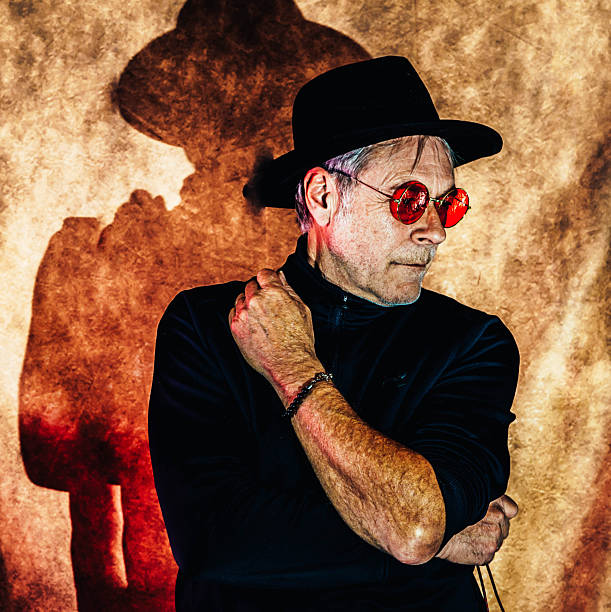 Artistic portrait of a man wearing a fedora hat and red glasses casting a sharp shadow on a textured background. Dramatic lighting with warm tones and retro cinematic style.