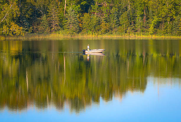 A lone fisherman stands in a small boat on a calm lake, surrounded by reflections of green forest along the shoreline. The tranquil scene conveys solitude and outdoor recreation in a natural setting.