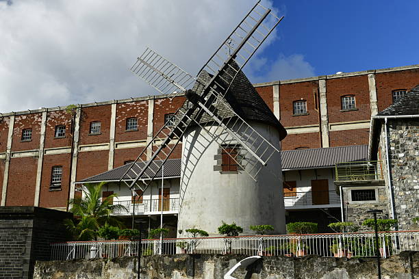 A pink wooden windmill stands in a simple structure, adorned with white metal ornaments on the roof and walls, situated in front of a green grassy park landscape. In the distance, an urban area building can be seen, surrounded by trees.