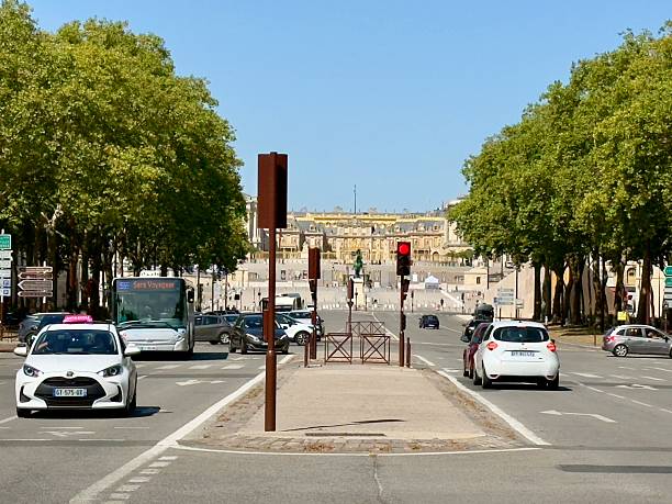 Horizontal cityscape photo of cars driving on the central road leading to the Palace of Versailles, seen in the distant background, on a sunny day in Summer