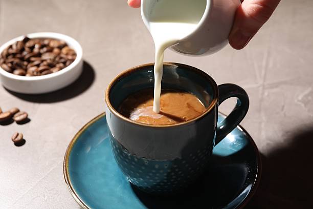 istock Woman pouring milk into cup with aromatic coffee at light table, closeup 2254706224