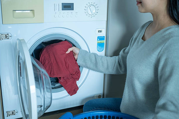 Closed-up of a woman is loading dirty clothes into a washing machine. Mature woman sits on the floor, placing clothes from a laundry basket into the machine while doing housework.
