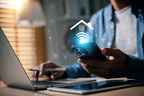istock Man smartphone to manage a smart home system via Wi-Fi connection, represented by a glowing house icon with wireless signals. He is also working on a laptop, symbolizing modern connectivity and remote control of home automation. 2254711723