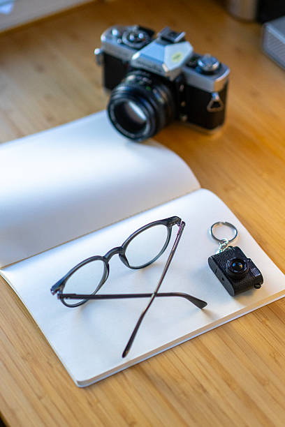 Vintage film camera, eyeglasses, and a small camera keychain placed on an open notebook on a wooden desk