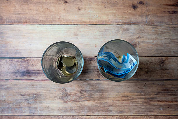 Top view of two glasses containing euro coins and euro banknotes on a wooden surface, illustrating financial inequality, savings disparity, and economic hardship. The image symbolizes personal finance challenges, cost of living issues, and income differences in a European context.
