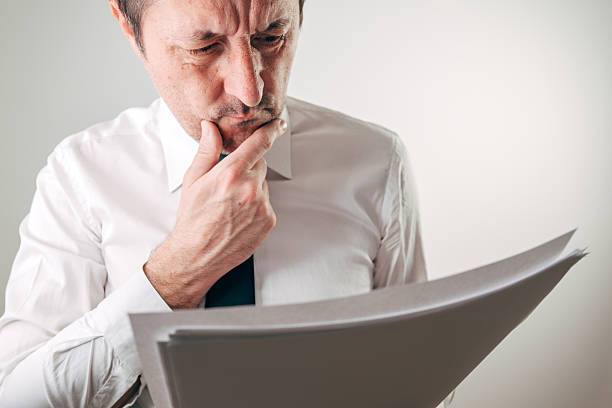 istock Businessman frowning deeply while holding and reading a thick stack of papers, hand thoughtfully on chin, for financial worry, bad news, contract review and overwork concepts. 2254713858