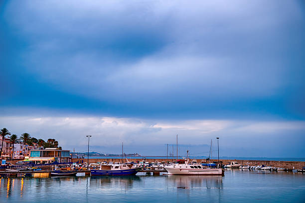 Boats are moored in a harbor under a cloudy sky. The scene shows the waterfront with a calm water surface reflecting the boats. People can be seen walking along the docks.