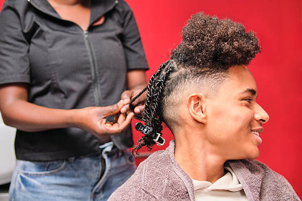 Afro man turns his head and smiles as the hairstylist braids his hair from behind, with only her hands visible.