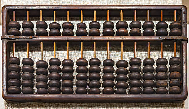 This full-frame, eye-level photograph showcases an antique Chinese abacus, known as a suanpan, meticulously crafted from dark wood. The abacus features a sturdy rectangular frame and a central crossbar, dividing the beads into two decks. Each of the thirteen vertical rods holds two upper beads (representing fives) and five lower beads (representing ones), a classic configuration for traditional Chinese calculations. The rich, deep brown tones of the wood, along with visible signs of wear and a subtle patina, suggest its age and long history of use. The textured, neutral-toned background, possibly a fabric, provides a gentle contrast, highlighting the abacus as a focal point and emphasizing its cultural and historical significance as a calculating tool.