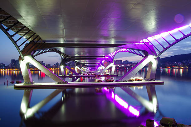 Low angle view looking underneath a modern bridge spanning across a river at night. The structure is illuminated by purple LED lights, creating geometric reflections on the water surface. The concept conveys urban infrastructure and connectivity. Authentic real photography.