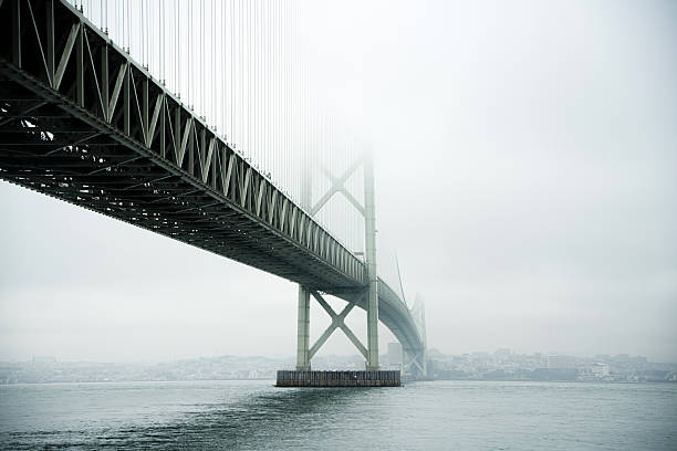 A wide-angle, low-angle shot of the Akashi Kaikyo Bridge spanning across the sea, partially disappearing into a thick, ethereal morning fog. The minimalist composition features a cold color palette, emphasizing a serene, mysterious, and peaceful atmosphere. The industrial structure of the bridge contrasts with the soft natural mist, creating a cinematic and dreamlike landscape in Hyogo, Japan.