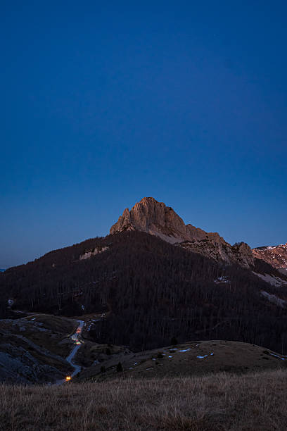 A snow-dusted mountain ridge illuminated by early sunrise light under a clear blue-hour sky. Dramatic, elongated shadows stretch across densely forested valleys and rugged limestone foreground, creating a layered alpine landscape with strong contrast, depth, and tranquil winter atmosphere.