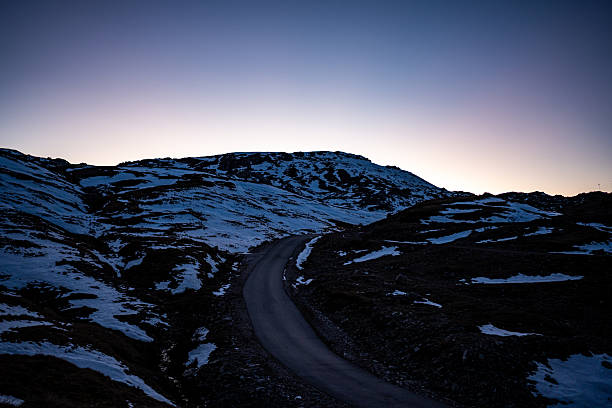 A snow-dusted mountain ridge illuminated by early sunrise light under a clear blue-hour sky. Dramatic, elongated shadows stretch across densely forested valleys and rugged limestone foreground, creating a layered alpine landscape with strong contrast, depth, and tranquil winter atmosphere.