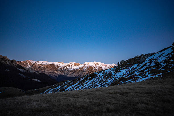 A snow-dusted mountain ridge illuminated by early sunrise light under a clear blue-hour sky. Dramatic, elongated shadows stretch across densely forested valleys and rugged limestone foreground, creating a layered alpine landscape with strong contrast, depth, and tranquil winter atmosphere.