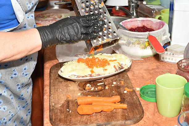 A woman's gloved hands shred boiled carrots using a hand-held metal grater.