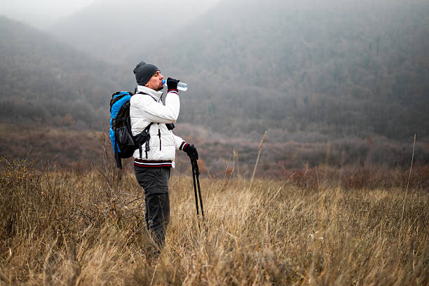 Hiker in warm clothing with backpack and hiking poles pausing to rest and drink water during hiking on foggy winter day.
