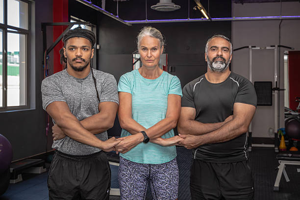 A middle-aged white woman, a young multiracial man, and an older male trainer stand side by side with interlocked hands, symbolizing unity, teamwork, and multigenerational fitness collaboration