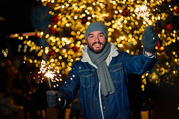 Smiling young adult man wearing winter clothes holding sparklers, celebrating Christmas or New Year's Eve against a blurry background of golden bokeh lights