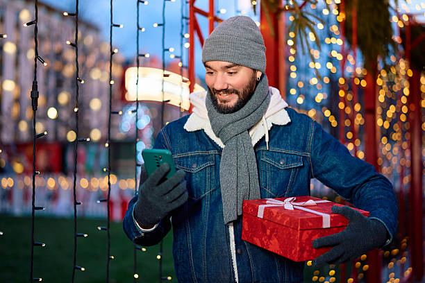 istock Smiling man holding gift box and using smartphone during holidays 2254752371