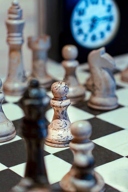 A chessboard and white and black chess pieces for playing chess. sixteen pieces : one king, one queen, two rooks, two bishops, two knights, and eight pawns. clock timer in the background.