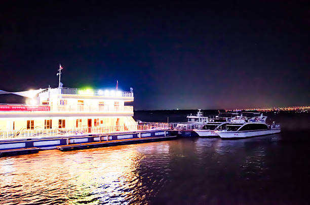 Night view of a brightly lit yellow cruise boat and several yachts moored along a calm river, with colorful reflections shimmering on the water surface.