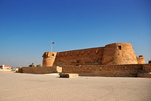 Arad, Bahrain: Arad Fort on Muharraq Island, showing the square defensive perimeter and the open space surrounding the structure. The fort, an example of Omani and Islamic military architecture from the 15th and 16th centuries, was restored in the 1980s using authentic materials like coral stone and lime. The image captures the fort’s low-profile defensive walls and one of its four cylindrical watchtowers under a clear sky.