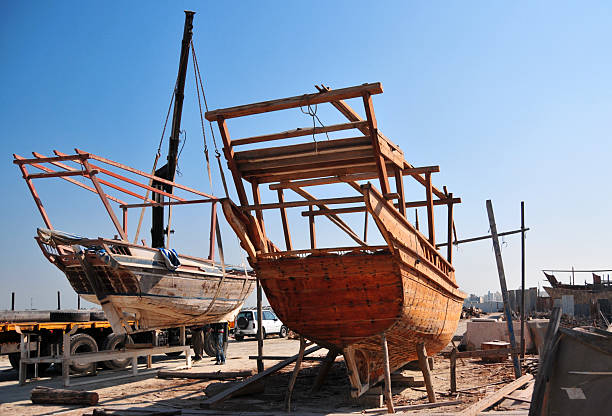 Muharraq Island, Bahrain: a dhow shipyard on the coast of Muharraq, illustrating the ongoing tradition of wooden boatbuilding. The image captures the skeletal wooden frames of traditional sailing vessels (dhows) under construction or repair. These vessels have historically been central to Bahrain’s maritime economy, including pearling and regional trade. The shipyard combines traditional craftsmanship with modern industrial equipment, such as cranes and transport vehicles, situated against the backdrop of the Muharraq shoreline and the Persian Gulf.