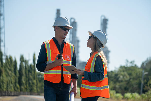 istock Industrial engineer wearing safety helmets and vests inspection technical plan and data on tablet with tall refinery towers rise background is industrial safety and carbon credit environment operations concept. 2254762601