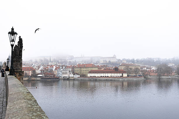 Prague, Czech republic - December 18, 2025: charles bridge statues and lamp post overlooking misty vltava and prague castle in winter fog