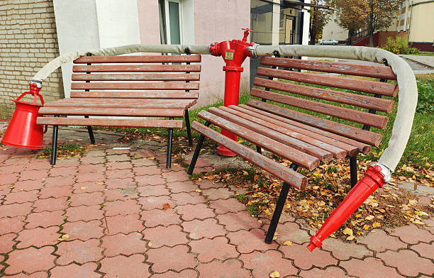 Red fire hydrant stands between two wooden benches, a fire hose draped over backrests. Red metal bucket hangs from left bench. Pavement path with autumn leaves, green grass, building wall in background.