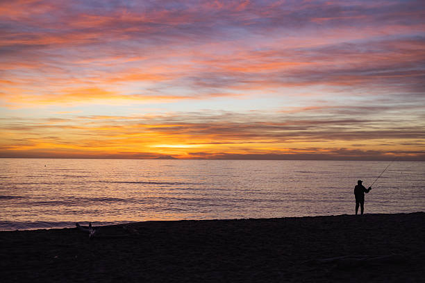 Silhouette of a lone fisherman casting a line along the sandy coast of Alberese at sunset, with calm sea and colorful sky creating a quiet and atmospheric coastal scene.