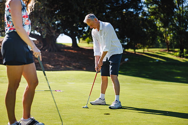 Senior Caucasian male golfer focusing carefully on aligning a putt on a well-maintained green course with a companion observing nearby during a sunny day.