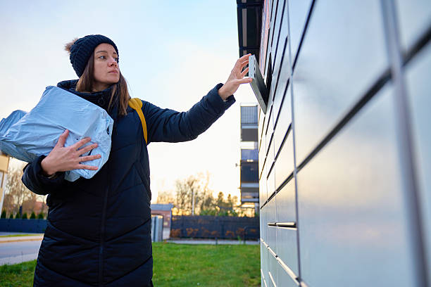 Woman standing outdoors holding package while using parcel locker touchscreen to collect delivery. Concept of online shopping